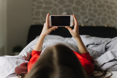 Pretty teenage girl using app on phone, talking on online video call, laughing, secretly watching movie, playing online games, lying under covers on bed. Generation alpha concept. Part of a seriesの写真素材