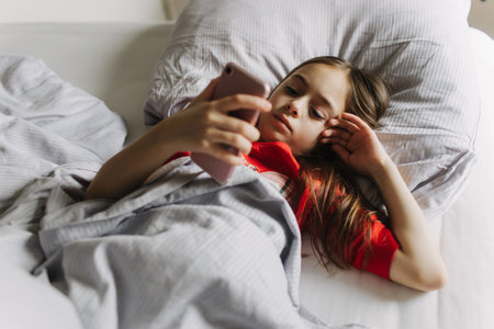 Pretty teenage girl using app on phone, talking on online video call, laughing, lying under covers...