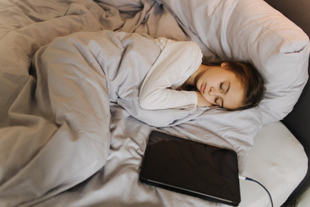 Pretty teenage girl fell asleep with a tablet in her hands, secretly watching movie, playing online games, lying under covers on bed. Generation alpha concept. Part of a seriesの写真素材