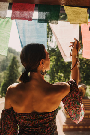 A person from behind holding prayer beads near colorful prayer flags in sunlight. The scene captures a peaceful spiritual moment outdoors.の写真素材