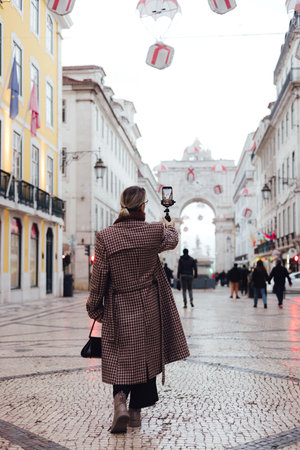 Vlogger traveler woman recording video on phone share via app her travel vacation in the Lisbon. Middle age woman influencer shooting social media stories on the street leading to Commerce Square. UGCの写真素材