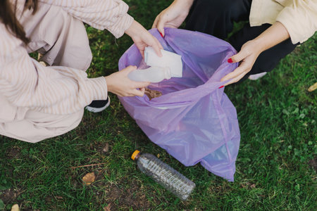 Close-up of mother and daughter's hands collecting waste in disposable trash bag on the public park. Volunteers help to keep nature clean up and pick up garbage. Environmental cleanup concept.の写真素材
