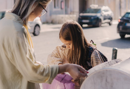Mom teaching Gen Alpha girl to sort trash near waste bins outdoors. Throwing plastic, paper, glass, e-waste bag into bucket for reuse. Concept of helping climate change and clean eco green no wasteの写真素材