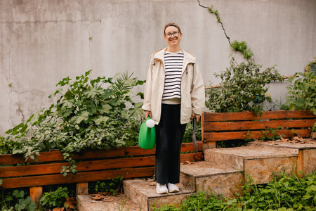 Portrait of a middle-aged woman as florist taking care of flowers and plants in garden with vintage greenhouse eco-farm. Concept green tourism, slow life, gardening, eco friendly. Part of the seriesの写真素材