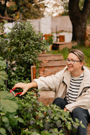 Portrait of a middle-aged woman as florist taking care of flowers and plants in garden with vintage greenhouse eco-farm. Concept green tourism, slow life, gardening, eco friendly. Part of the seriesの写真素材