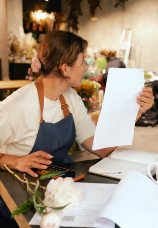 A middle-aged woman calculates finances on a calculator at a flower shop with various plants in the...