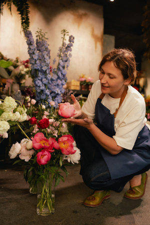Middle-aged female florist in apron arranging fresh flowers for bouquet in the flower shop. Small...