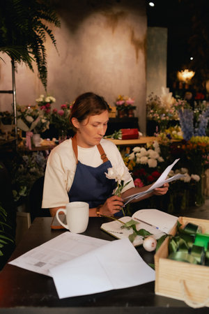 A middle-aged woman calculates finances on a calculator at a flower shop with various plants in the...