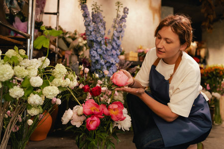 Middle-aged female florist in apron arranging fresh flowers for bouquet in the flower shop. Small local business. Part of a series. Soft focusの写真素材