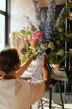 Middle-aged female florist in apron arranging fresh flowers for bouquet She ties a ribbon on the bouquet in the flower shop. Small local business. Part of a series. Soft focus.の写真素材