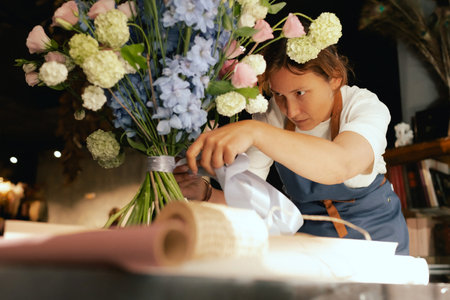Middle-aged female florist in apron arranging fresh flowers for bouquet She ties a ribbon on the bouquet in the flower shop. Small local business. Part of a series. Soft focus.の写真素材