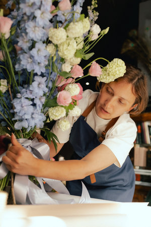 Middle-aged female florist in apron arranging fresh flowers for bouquet She ties a ribbon on the bouquet in the flower shop. Small local business. Part of a series. Soft focus.の写真素材