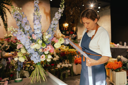 Middle-aged female florist in apron arranging fresh flowers for bouquet She ties a ribbon on the bouquet in the flower shop. Small local business. Part of a series. Soft focus.の写真素材