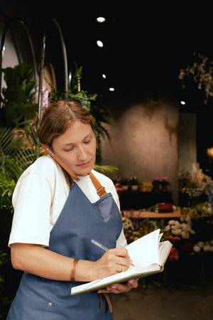 Middle-aged female florist in apron arranging flowers. She is arranging logistics, and delivery, taking orders. Small local business. Part of a series. Soft focusの写真素材