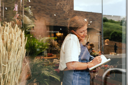 Middle-aged female florist in apron arranging logistics and delivery, taking orders. Small local business. Part of a series. Soft focusの写真素材