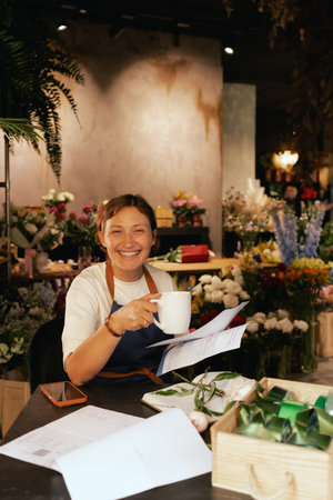 A middle-aged woman calculates finances on a calculator at a flower shop with various plants in the background. She is arranging logistics, and delivery, taking orders. Small local businessの写真素材