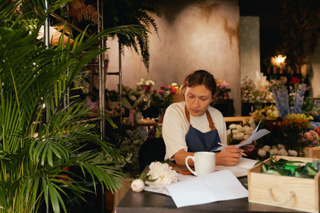 A middle-aged woman calculates finances on a calculator at a flower shop with various plants in the background. She is arranging logistics, and delivery, taking orders. Small local businessの写真素材