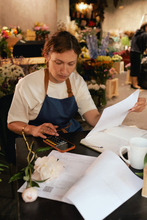 A middle-aged woman calculates finances on a calculator at a flower shop with various plants in the background. She is arranging logistics, and delivery, taking orders. Small local businessの写真素材