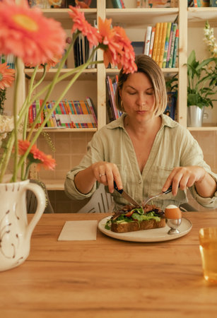 Crop middle-aged woman wearing stylish outfit eating delicious breakfast including egg and bacon with freshly greens served on wooden table in sunny outdoor cafe. Part of a series.の写真素材