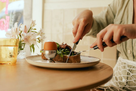 Close-up of female hands with cutlery having breakfast with egg and bacon with freshly greens served on wooden table in sunny outdoor cafe. Part of a series.の写真素材
