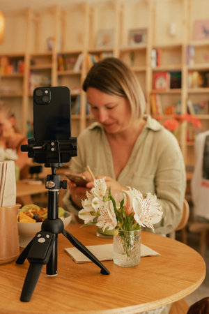 Middle-aged woman is video blogging in a cafe on a phone with a tripod. Small business. Using phone for business, online shopping, transfer money, financial, internet banking. Part of a series.の写真素材