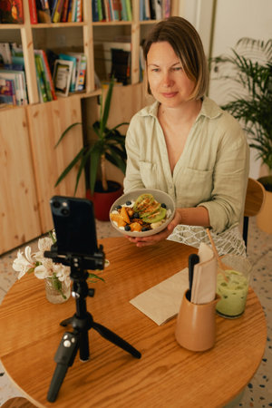 Middle-aged woman is video blogging in a cafe on a phone with a tripod. Small business. Using phone for business, online shopping, transfer money, financial, internet banking. Part of a series.の写真素材