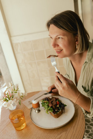 Crop middle-aged woman wearing stylish outfit eating delicious breakfast including egg and bacon with freshly greens served on wooden table in sunny outdoor cafe. Part of a series.の写真素材