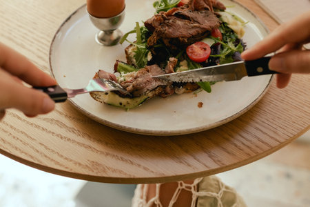 Close-up of female hands with cutlery having breakfast with egg and bacon with freshly greens served on wooden table in sunny outdoor cafe. Part of a series.の写真素材