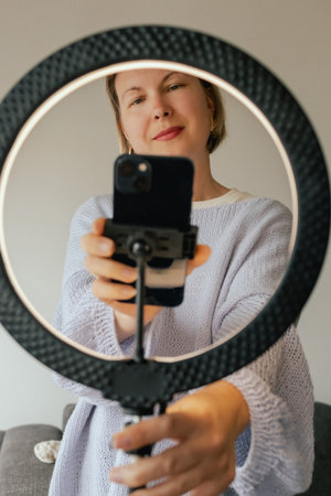 Middle-aged woman beauty blogger filming skincare product unboxing at home with ring light. User-generated content UGC for online store promotion. Vlogger live streaming. Part of a seriesの写真素材