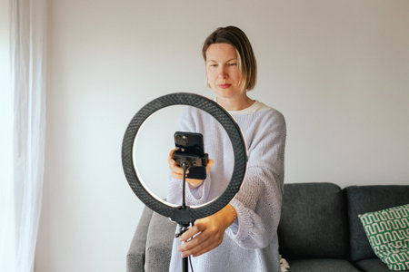 Middle-aged woman beauty blogger filming skincare product unboxing at home with ring light. User-generated content UGC for online store promotion. Vlogger live streaming. Part of a seriesの写真素材