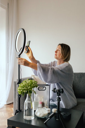 Middle-aged woman beauty blogger filming skincare product unboxing at home with ring light. User-generated content UGC for online store promotion. Vlogger live streaming. Part of a seriesの写真素材