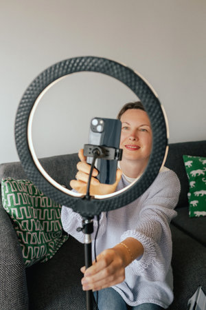 Middle-aged woman beauty blogger filming skincare product unboxing at home with ring light. User-generated content UGC for online store promotion. Vlogger live streaming. Part of a seriesの写真素材