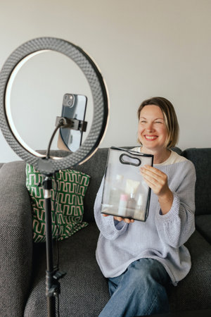 Middle-aged woman beauty blogger filming skincare product unboxing at home with ring light. User-generated content UGC for online store promotion. Vlogger live streaming. Part of a seriesの写真素材