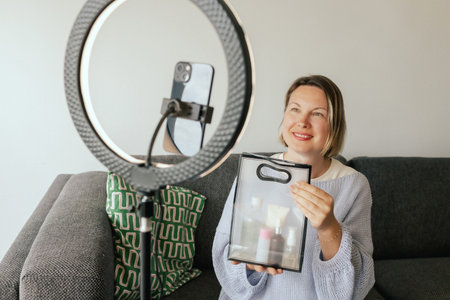 Middle-aged woman beauty blogger filming skincare product unboxing at home with ring light....