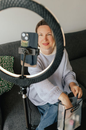Middle-aged woman beauty blogger filming skincare product unboxing at home with ring light. User-generated content UGC for online store promotion. Vlogger live streaming. Part of a seriesの写真素材
