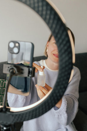 Middle-aged woman beauty blogger filming skincare product unboxing at home with ring light. User-generated content UGC for online store promotion. Vlogger live streaming. Part of a seriesの写真素材