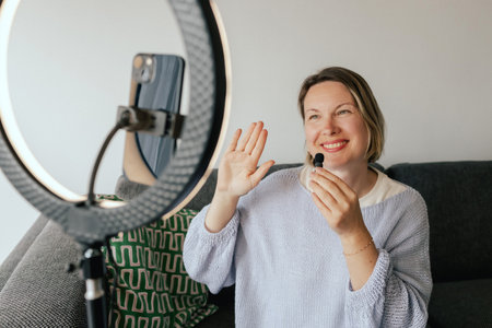 Middle-aged woman beauty blogger filming skincare product unboxing at home with ring light. User-generated content UGC for online store promotion. Vlogger live streaming. Part of a seriesの写真素材