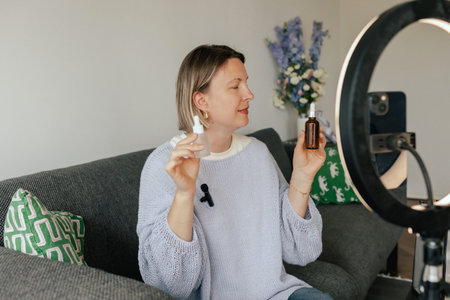 Middle-aged woman beauty blogger filming skincare product unboxing at home with ring light. User-generated content UGC for online store promotion. Vlogger live streaming. Part of a seriesの写真素材