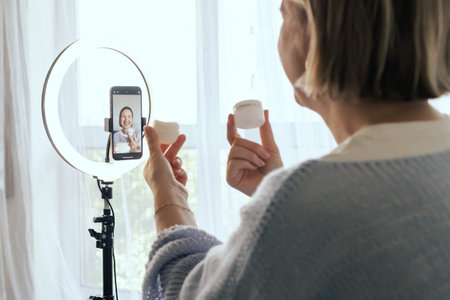 Middle-aged woman beauty blogger filming skincare product unboxing at home with ring light. User-generated content UGC for online store promotion. Vlogger live streaming. Part of a seriesの写真素材