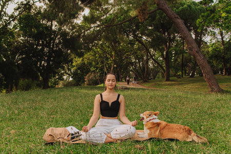 Fit sporty mindful young woman meditating, doing yoga and breathing exercises with eyes closed, feeling peace and balance in tropical park with her Corgi dog on a sunny day.の写真素材