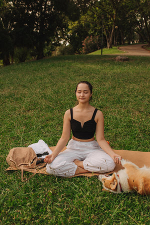 Fit sporty mindful young woman meditating, doing yoga and breathing exercises with eyes closed, feeling peace and balance in tropical park with her Corgi dog on a sunny day.の写真素材