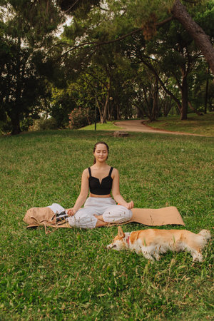 Fit sporty mindful young woman meditating, doing yoga and breathing exercises with eyes closed, feeling peace and balance in tropical park with her Corgi dog on a sunny day.の写真素材