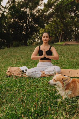Fit sporty mindful young woman meditating, doing yoga and breathing exercises with eyes closed, feeling peace and balance in tropical park with her Corgi dog on a sunny day.の写真素材