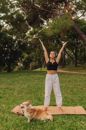 Fit sporty mindful young woman meditating, doing yoga and breathing exercises with eyes closed, feeling peace and balance in tropical park with her Corgi dog on a sunny day.の写真素材