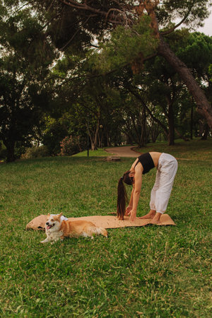 Fit sporty mindful young woman meditating, doing yoga and breathing exercises with eyes closed, feeling peace and balance in tropical park with her Corgi dog on a sunny day.の写真素材