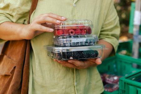 Close-up of hand with fresh berry in plastic packaging at local urban market. Organic produce on...