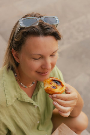 Gen Z middle age woman tries Portuguese egg tart pastry called Pastel de Nata near bakery in Lisbon....