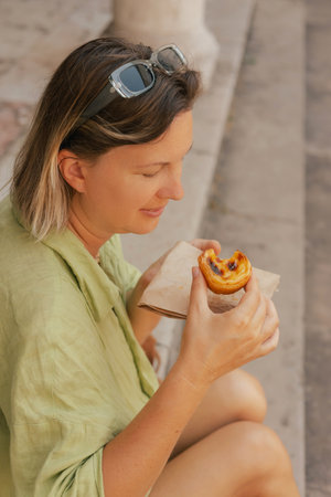 Gen Z middle age woman tries Portuguese egg tart pastry called Pastel de Nata near bakery in Lisbon....