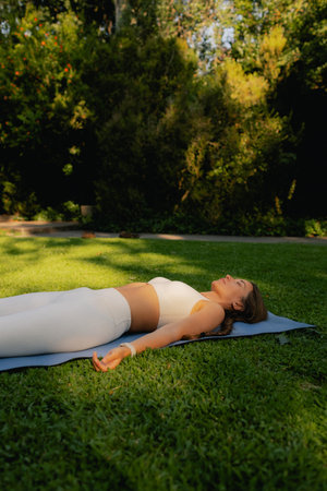Young woman in sportswear stretching body on fitness mat at public park, doing yoga. Healthy lifestyle, sport. Fitness. Girl doing yoga in morning park. Part of a series. Soft focus.の写真素材