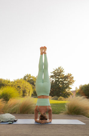 Young woman in sportswear standing on her head on fitness mat at public park, doing yoga. Healthy...